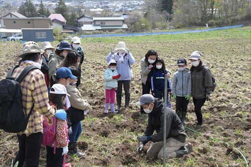 山田さんがブドウの苗木の植え方を説明、説明を聞く参加者一同